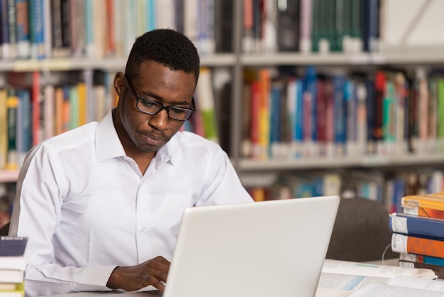 In de bibliotheek knappe Afrikaanse mannelijke student met laptop en boeken werken in een middelbare school Universiteitsbibliotheek ondiepe scherptediepte