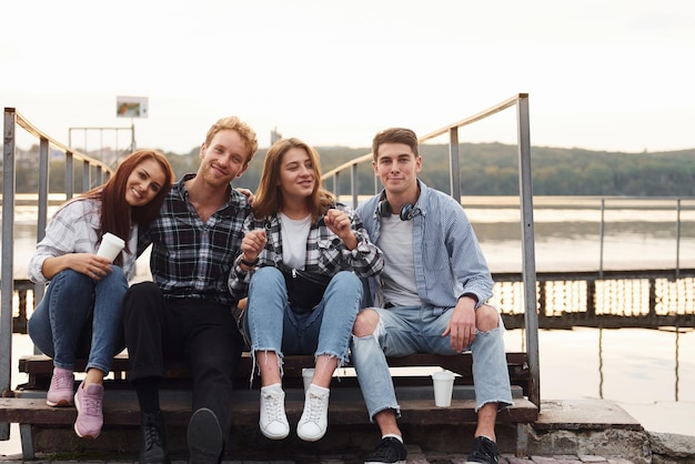 In de buurt van het meer zitten en elkaar omhelzen Groep jonge vrolijke vrienden die buiten samen plezier hebben