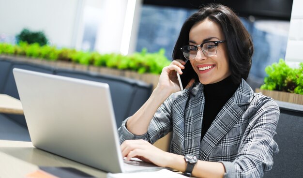 In de coworking. Zijaanzicht foto van een vrolijke vrouw in slimme vrijetijdskleding, pratend aan de telefoon terwijl ze op haar laptop werkt.