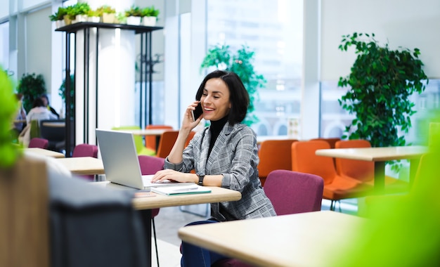 In de coworking. Zijaanzicht foto van een vrolijke vrouw in slimme vrijetijdskleding, pratend aan de telefoon terwijl ze op haar laptop werkt.