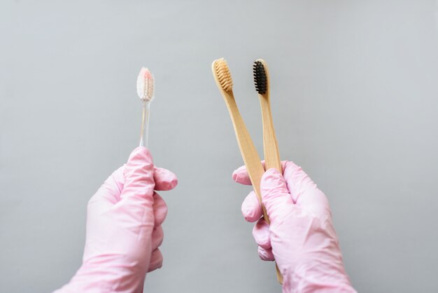 In de ene hand een kunststof tandenborstel, in de andere twee bamboe tandenborstels. Handen in roze handschoenen houden borstels.