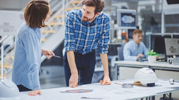 In de Industrial Engineering Facility werkt jonge vrouwelijke ontwerper met Chief Industrial Engineer Ze hebben discussie Analyse en Correcte Engine Design Technische Drafts die op de tafel liggen