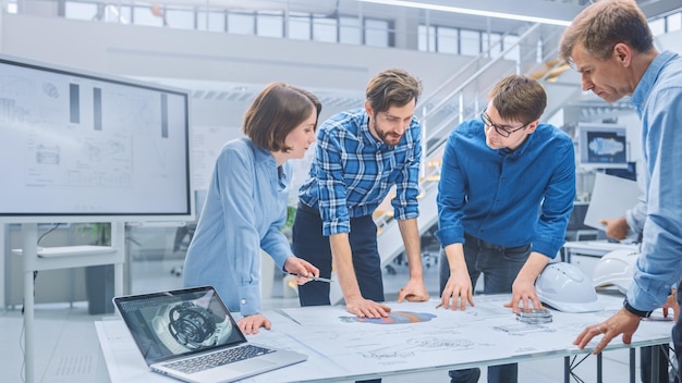 In de industriële ingenieursfaciliteit verzamelt een diverse groep ingenieurs en technici zich rond een tafel met technische ontwerpen van motoren, bespreken ze technologieën en analyseren ze.