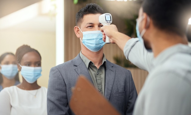 In de rij om gescreend te worden bijgesneden opname van een knappe, volwassen zakenman met een masker en zijn temperatuur gemeten terwijl hij aan het hoofd van een rij in zijn kantoor staat