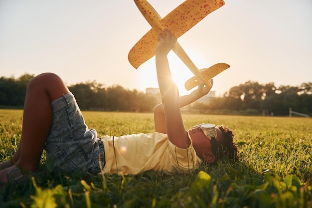 In een bril met speelgoedvliegtuig heeft Afro-Amerikaanse jongen plezier in het veld op zomerdag