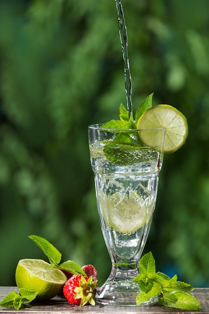 In een hoog glas met ijs en limoen stroomt een stroom water, daarna een aardbei, schijfjes limoen en muntblad