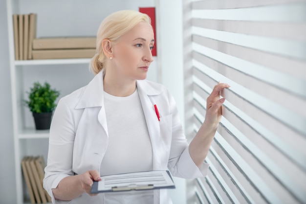 In gedachten verzonken. Vrouw met blond haar in een witte jas met een map in haar hand staan in de buurt van het raam.