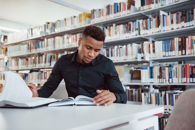 In stijlvol zwart overhemd. afro-amerikaanse man zit in de bibliotheek en op zoek naar wat informatie in de boeken