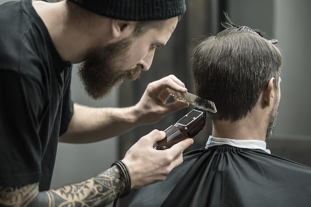 Incomparable barber with a beard and a tattoo is cutting the hair of his client in the black cape in the barbershop