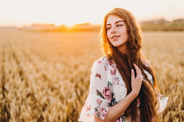 Фото Невероятная молодая женщина с длинными вьющимися волосами и веснушками лицом. женщина в платье позирует в пшеничном поле на закате. портрет близкий вверх