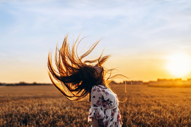 Невероятная молодая женщина с длинными вьющимися волосами. женщина в платье позирует на пшеничном поле на закате и выпрямляет голову