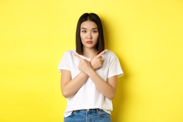 Indecisive asian girl need help with choice, pointing fingers sideways and looking confused, standing over yellow background.