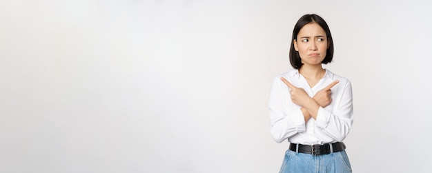 Indecisive asian woman pointing fingers sideways pointing fingers and looking clueless confused with choices standing over white background