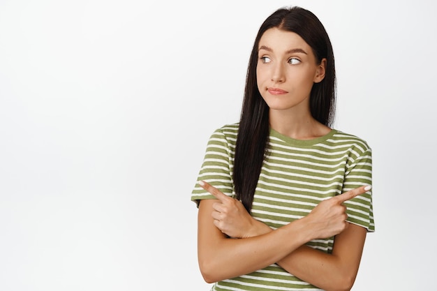 Foto cliente ragazza indecisa che punta le dita lateralmente e guardando a sinistra con un'espressione del viso premurosa che prende una decisione in piedi su sfondo bianco