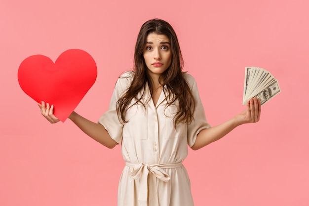 Indecisive and unsure, cute lovely modern woman cant decide what important, shrugging looking confused, holding heart card and money, don't know whats right, questioned over pink wall