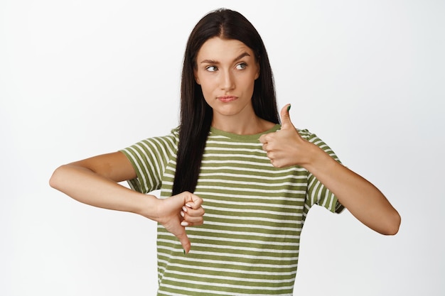 Indecisive woman showing thumbs up and thumbs down like or dislike making her feedback standing over white background