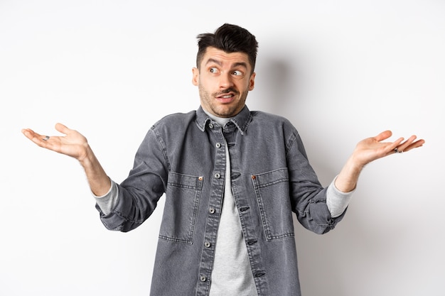 Indecisive young man looking aside and shrugging, know nothing, have no idea, standing clueless and confused against white background.