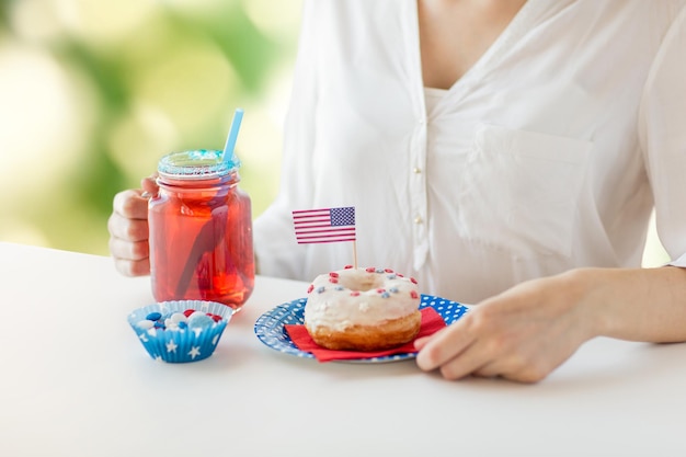 Giorno dell'indipendenza, celebrazione, patriottismo e concetto di vacanze - primo piano di una donna che mangia una ciambella glassata, beve succo da un grande barattolo di vetro e celebra il 4 luglio su sfondo naturale verde