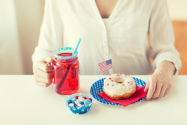 independence day, celebration, patriotism and holidays concept - close up of woman eating glazed sweet donut, drinking juice from big glass mason jar or mug and celebrating 4th july at home party