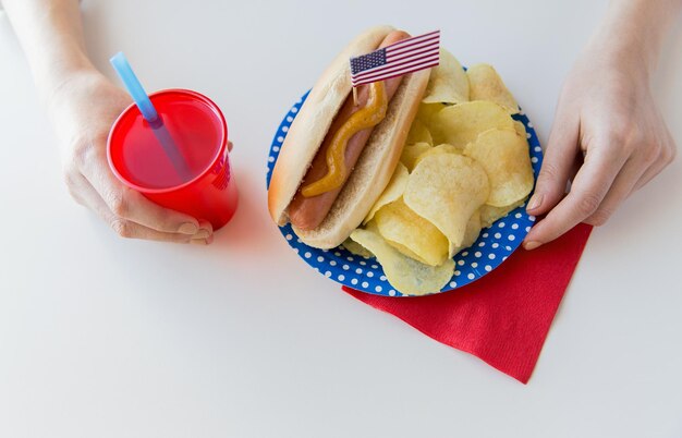 independence day, celebration, patriotism and holidays concept - close up of woman hands eating hot dog with american flag decoration, potato chips and juice in cup on 4th july at home party