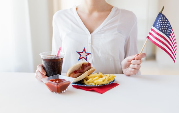 independence day, celebration, patriotism and holidays concept - close up of woman hands with hot dog and french fries holding american flag and coca cola drink on 4th july at home party