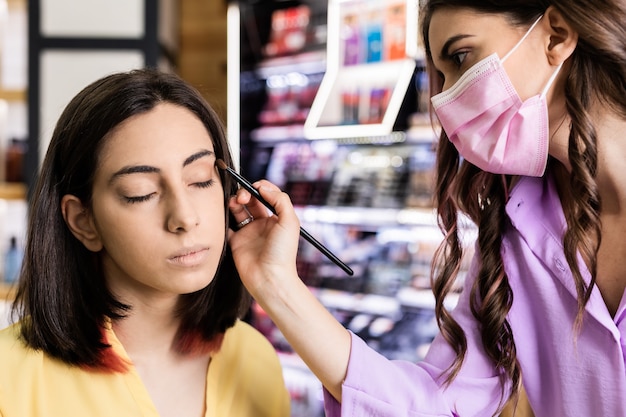 Independent makeup artist applying cosmetics to a young hispanic woman protected with medical mask for covid19 Lifestyle small business skin professional Yellow and lavender year color