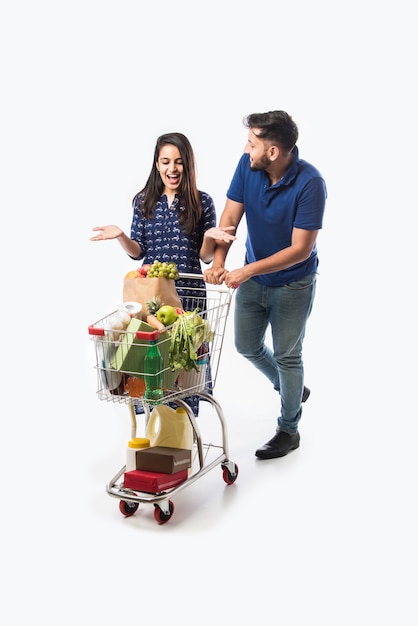 Indiaas jong stel met winkelwagentje of karretje vol met kruidenierswaren, groenten en fruit. Geïsoleerde foto van volledige lengte over witte muur