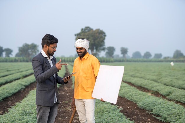 Indian agronomist or officer with farmer at agriculture field holding whiteboard