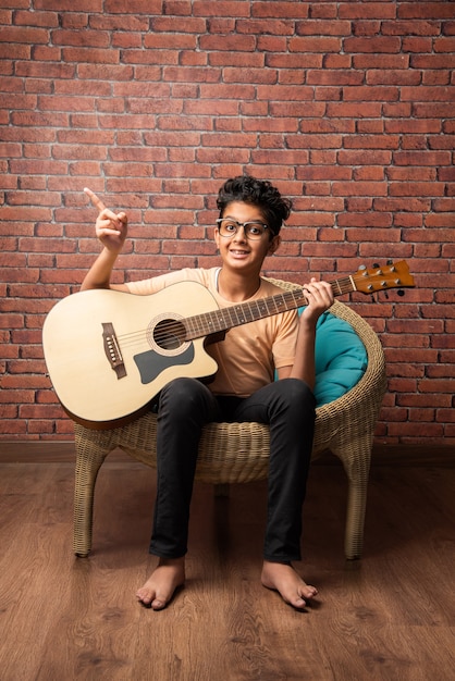 Indian asian boy playing acoustic  guitar while sitting against white wall 