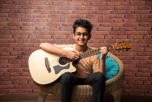 흰 벽에 앉아 어쿠스틱 기타를 연주하는 인도 아시아 소년