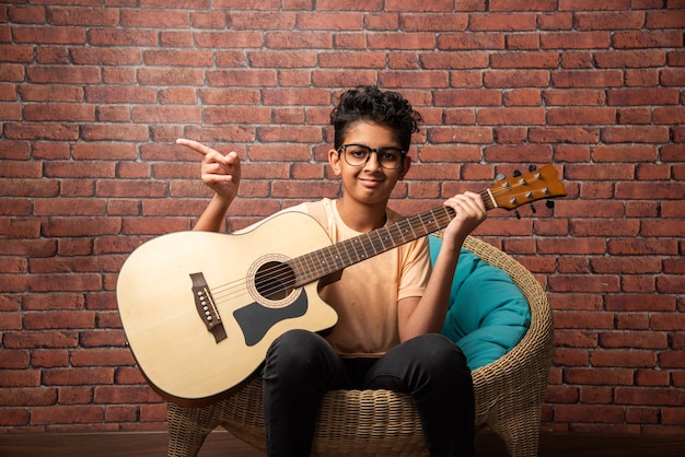 Indian asian boy playing acoustic  guitar while sitting against white wall 