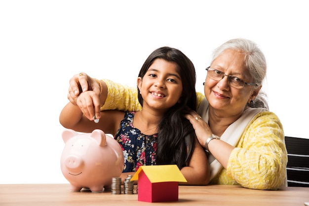 Indian or asian Grandma teaching cute grand daughter importance of savings, putting coins into pink piggy bank