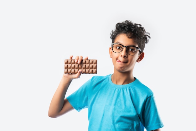 Indian asian happy boy eating chocolate slab, standing isolated against white wall