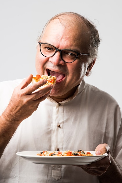 Indian asian senior or old man eating pizza while standing isolated against white wall