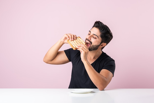 Photo indian asian young bearded man eats bread sandwich while sitting in kitchen or dining table. showing or presenting