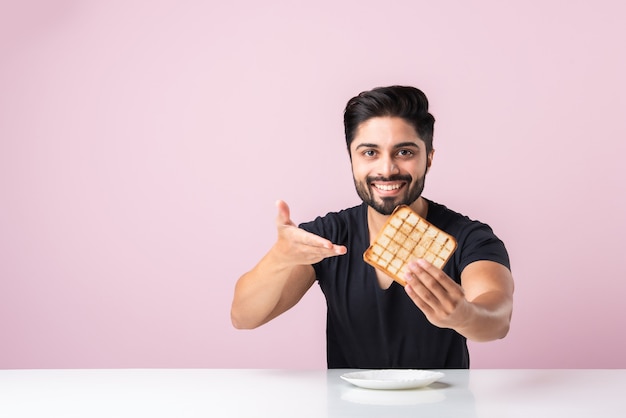 Indian Asian young bearded man eats bread sandwich while sitting in kitchen or dining table. Showing or presenting