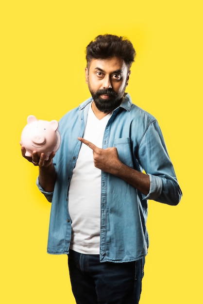 Indian asian young bearded man holding piggy bank on yellow. people and finance concept