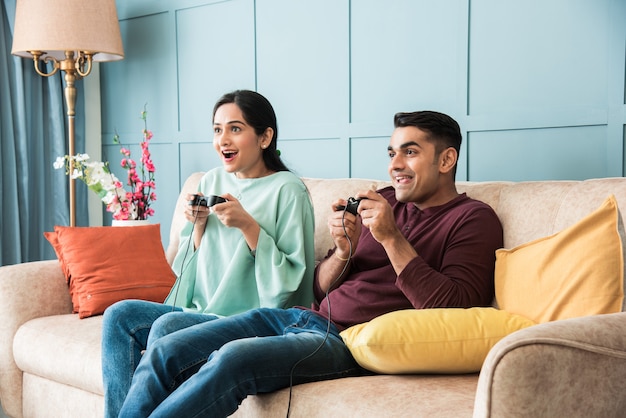 Indian asian young couple playing video game using joystick or\
controller while sitting on couch or sofa