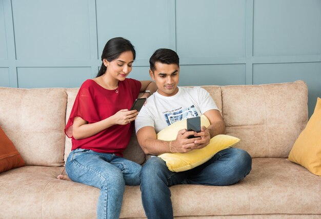 Indian asian young couple using smartphone together while sitting on sofa or couch and modern interior space or home