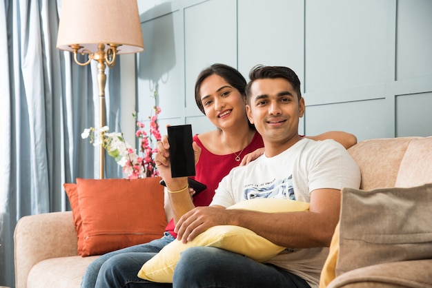 Indian asian young couple using smartphone together while sitting on sofa or couch and modern interior space or home