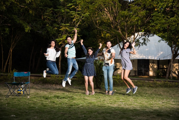 Indian asian young friends dancing or jumping while camping near tent, outdoors in jungle