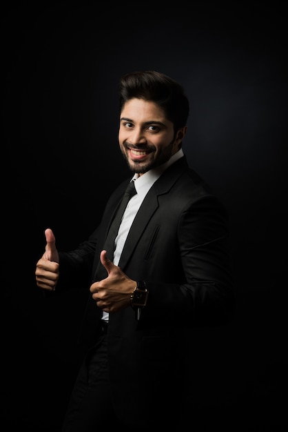 Photo indian bearded male businessman celebrating success with thumbs up or raising fist while standing isolated over black background