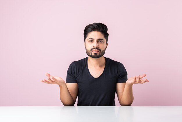 Indian bearded young man thinking while sitting isolated against pink background at table or desk