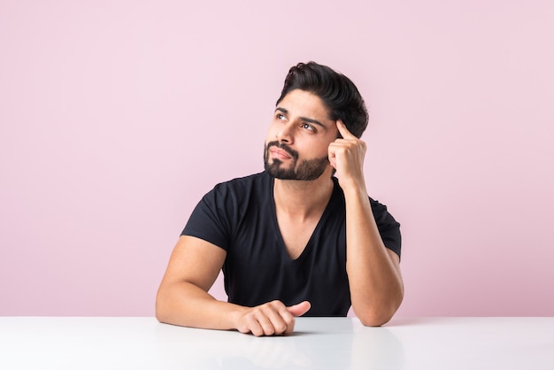 Indian bearded young man thinking while sitting isolated against pink background at table or desk