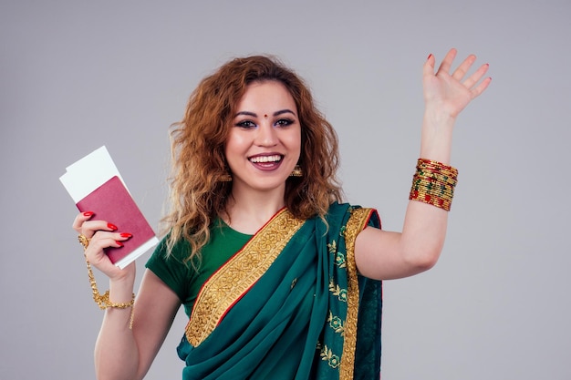 Indian beautiful fashion brunette woman in green traditional wedding rich sari with gold jewelry set professional makeup on white studio background bindi holding a passport and boarding passes