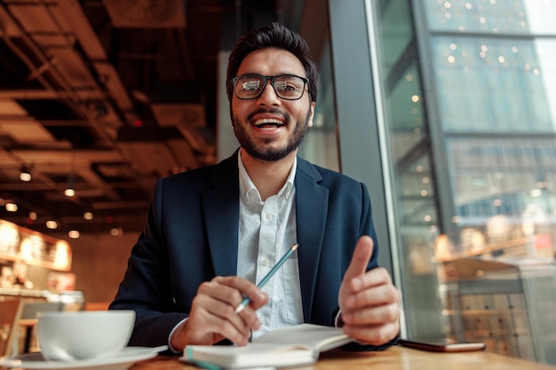 indian businessman is making notes in notepad during working day in cafe and looking at camera