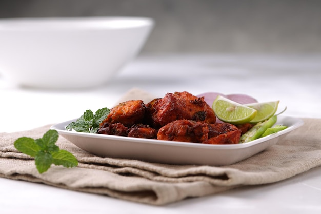 인도 치킨 튀김은 레몬 슬라이스로 장식된 녹색 칠리와 민트 잎으로 장식된 흰색 접시에 배열되어 회색 질감 배경 선택 초점이 있는 흰색 표면에 배치됩니다.