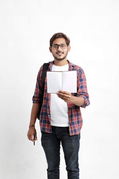 Indian college student holding schoolbag and showing note book on white wall