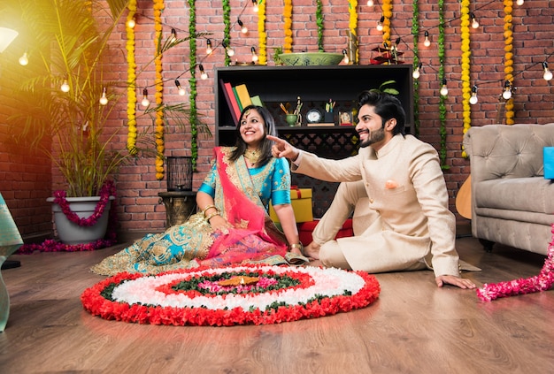 Indian couple making flower Rangoli on Diwali or Onam Festival, taking selfie or holding sweets