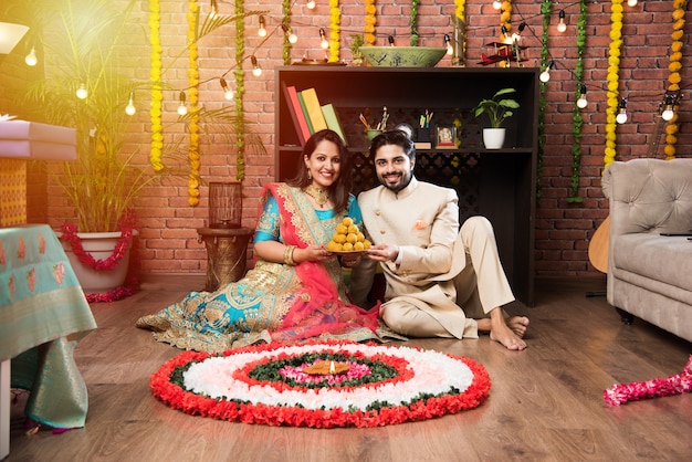 Indian couple making flower Rangoli on Diwali or Onam Festival, taking selfie or holding sweets